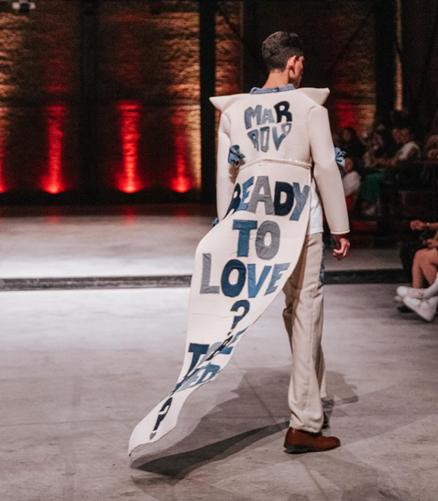 A male model on a fashion runway wearing a structured white coat with an elongated train reading MARROLO READY TO LOVE, perfectly illustrating how haute couture meets streetwear