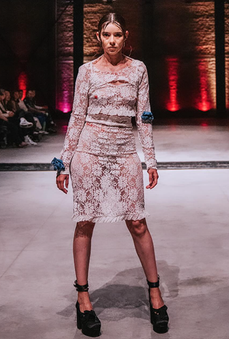 A female model on a fashion runway wearing a white lace two-piece garment with blue flower accents and black platform heels, showing how haute couture meets streetwear
